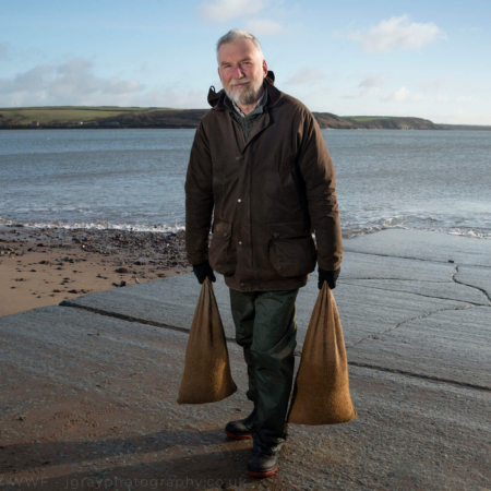 Mike Furness is standing on a slipway. He is carrying a hessian bag in each arm.